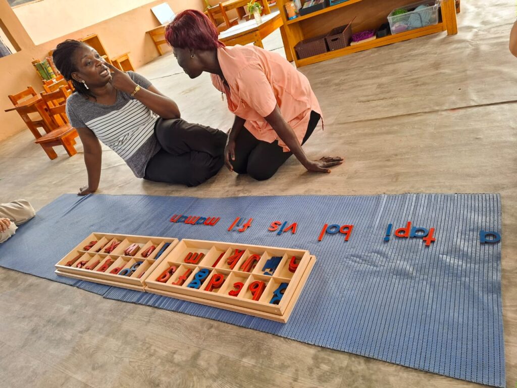 Two women sit on the floor by a blue mat with Montessori letters and a wooden tray in a classroom setting.